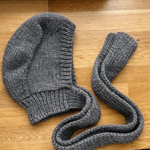 Gray knit hood laid flat on a wooden floor, showing the curved hood shape, textured stitches, and ribbed neckline; long knitted ties are laid beside it with one end folded over.