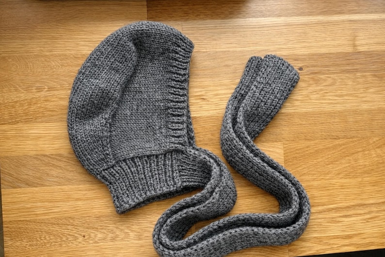 Gray knit hood laid flat on a wooden floor, showing the curved hood shape, textured stitches, and ribbed neckline; long knitted ties are laid beside it with one end folded over.
