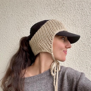 Op de afbeelding: Een zwarte honkbalpet met een beige gebreid oorwarmer-opzetstuk dat de zijkanten en de achterkant bedekt. De oorwarmer is gemaakt met een geribbelde textuur en bevat twee lange, bijpassende beige gebreide koorden die naar beneden hangen.