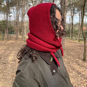 May include: A deep red knitted hood with a textured garter stitch pattern that covers the head and neck, featuring a thick folded collar section and two long knitted ties hanging down from the front to secure the garment around the chin.