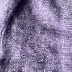 A soft, lavender fuzzy knitted bonnet lying on a light-colored wooden table. The bonnet features long matching ties and a fluffy, mohair-style texture. Bright, natural sunlight streams across the scene, highlighting the delicate fibers of the knitwear.