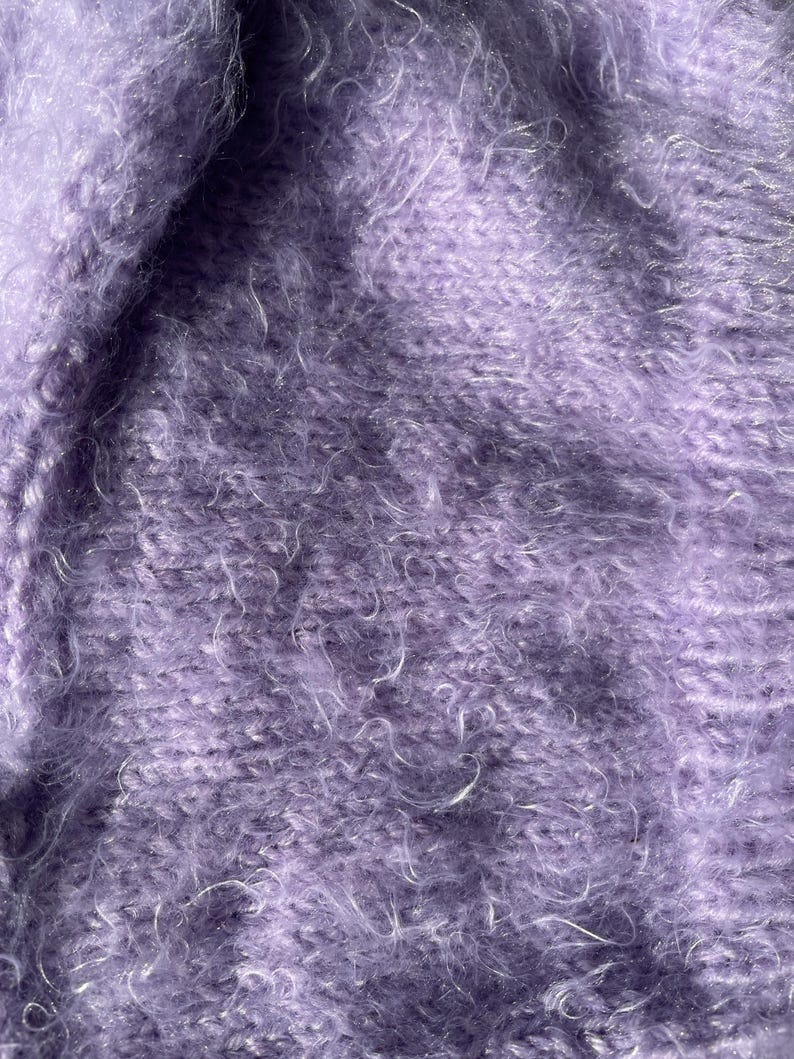 A soft, lavender fuzzy knitted bonnet lying on a light-colored wooden table. The bonnet features long matching ties and a fluffy, mohair-style texture. Bright, natural sunlight streams across the scene, highlighting the delicate fibers of the knitwear.