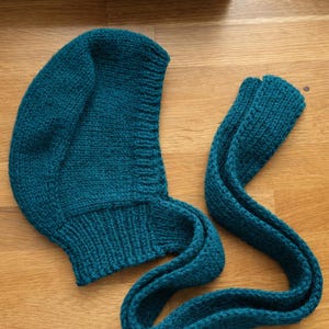 Teal knit hood laid flat on a wooden floor, showing the curved hood shape and ribbed neckline; a long knitted tie strap extends to the right with the end folded over.