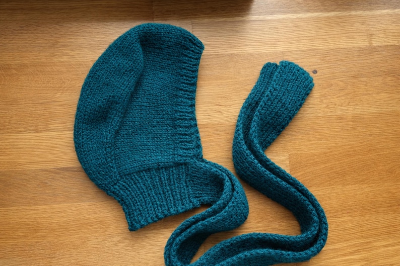Teal knit hood laid flat on a wooden floor, showing the curved hood shape and ribbed neckline; a long knitted tie strap extends to the right with the end folded over.