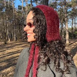 May include: A burgundy-colored knitted hood with long ties, worn over dark curly hair. The hood is made of a fuzzy material. The person is wearing a gray coat and sunglasses, with a forest background.