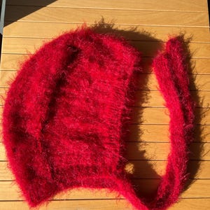A soft, red fuzzy knitted bonnet lying on a light-colored wooden table. The bonnet features long matching ties and a fluffy, mohair-style texture. Bright, natural sunlight streams across the scene, highlighting the delicate fibers of the knitwear.