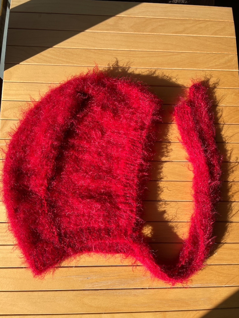 A soft, red fuzzy knitted bonnet lying on a light-colored wooden table. The bonnet features long matching ties and a fluffy, mohair-style texture. Bright, natural sunlight streams across the scene, highlighting the delicate fibers of the knitwear.