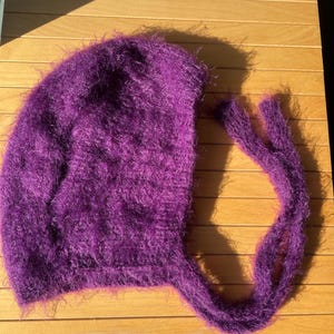 A soft, dark purple fuzzy knitted bonnet lying on a light-colored wooden table. The bonnet features long matching ties and a fluffy, mohair-style texture. Bright, natural sunlight streams across the scene, highlighting the delicate fibers of the knitwear.