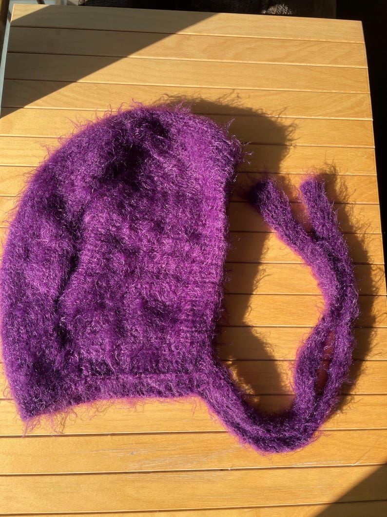 A soft, dark purple fuzzy knitted bonnet lying on a light-colored wooden table. The bonnet features long matching ties and a fluffy, mohair-style texture. Bright, natural sunlight streams across the scene, highlighting the delicate fibers of the knitwear.