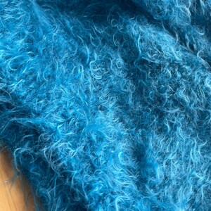 A soft, turquoise fuzzy knitted bonnet lying on a light-colored wooden table. The bonnet features long matching ties and a fluffy, mohair-style texture. Bright, natural sunlight streams across the scene, highlighting the delicate fibers of the knitwear.