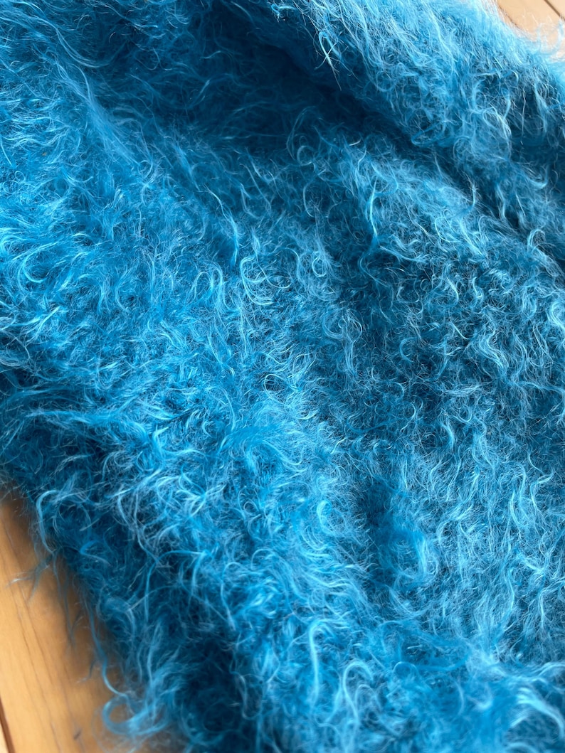 A soft, turquoise fuzzy knitted bonnet lying on a light-colored wooden table. The bonnet features long matching ties and a fluffy, mohair-style texture. Bright, natural sunlight streams across the scene, highlighting the delicate fibers of the knitwear.