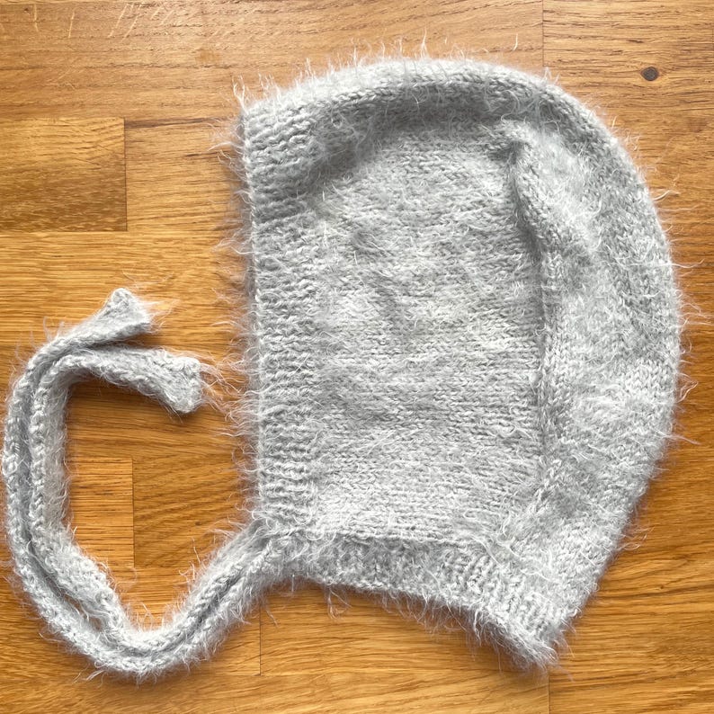 A soft, silver grey fuzzy knitted bonnet lying on a light-colored wooden table. The bonnet features long matching ties and a fluffy, mohair-style texture. Bright, natural sunlight streams across the scene, highlighting the delicate fibers of the knitwear.