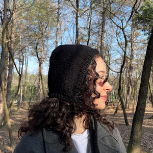 May include: A black knitted hood with long ties. The hood covers the head, ears, and neck. The person is wearing a grey coat and a white shirt. The background is a forest with tall trees and sunlight.