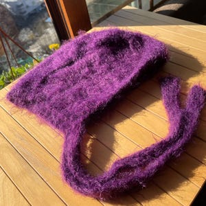 A soft, dark purple fuzzy knitted bonnet lying on a light-colored wooden table. The bonnet features long matching ties and a fluffy, mohair-style texture. Bright, natural sunlight streams across the scene, highlighting the delicate fibers of the knitwear.