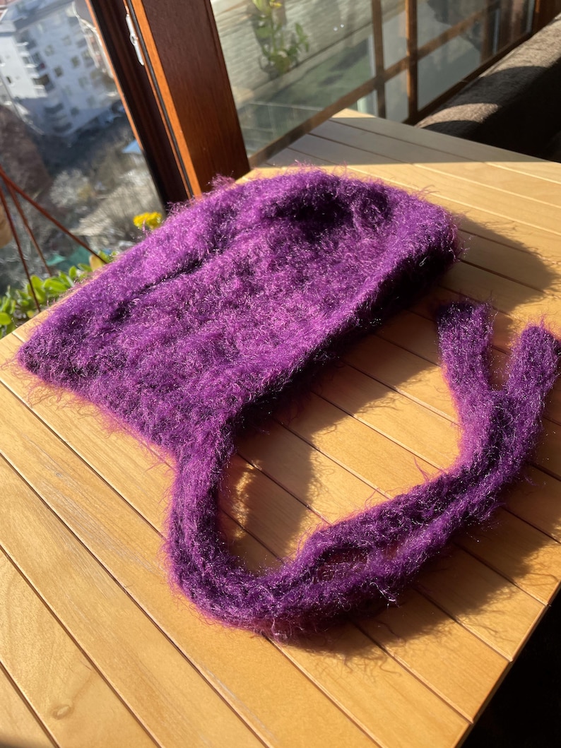 A soft, dark purple fuzzy knitted bonnet lying on a light-colored wooden table. The bonnet features long matching ties and a fluffy, mohair-style texture. Bright, natural sunlight streams across the scene, highlighting the delicate fibers of the knitwear.