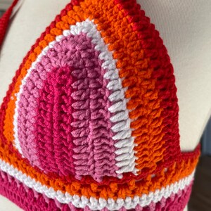 A close-up shot of a handmade crochet bikini top featuring the colors of the Lesbian Pride flag. The triangular cup is constructed with concentric rows of stitches in shades of dark pink, light pink, white, orange, and red. The vibrant, textured top is displayed on a white mannequin, highlighting the detailed yarn work and the horizontal striped pattern along the bottom band.