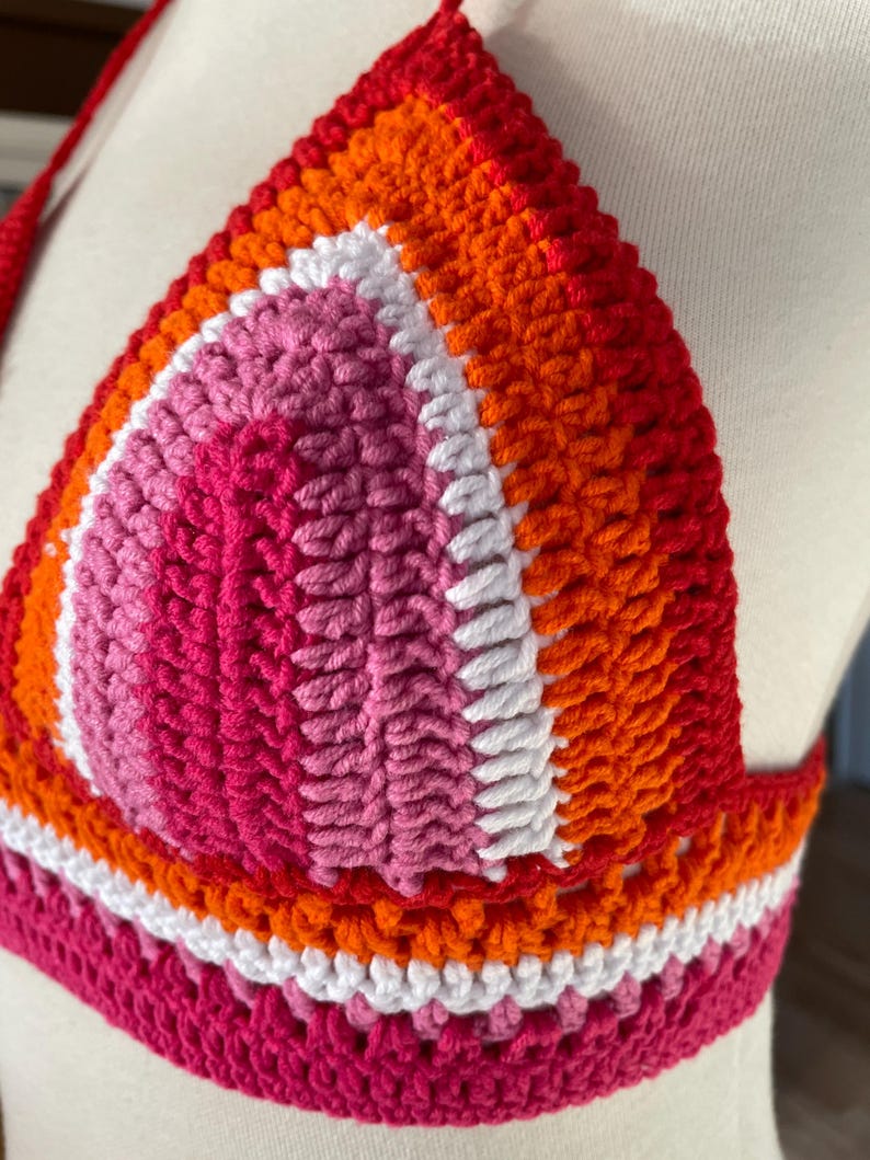 A close-up shot of a handmade crochet bikini top featuring the colors of the Lesbian Pride flag. The triangular cup is constructed with concentric rows of stitches in shades of dark pink, light pink, white, orange, and red. The vibrant, textured top is displayed on a white mannequin, highlighting the detailed yarn work and the horizontal striped pattern along the bottom band.