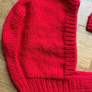 May include: A bright red hand-knitted bonnet featuring a textured garter stitch pattern on the main body and a ribbed knit border along the face opening. The headpiece includes a matching knitted strap extending from the bottom edge.