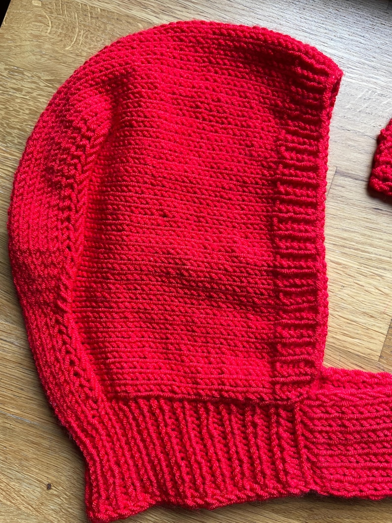 May include: A bright red hand-knitted bonnet featuring a textured garter stitch pattern on the main body and a ribbed knit border along the face opening. The headpiece includes a matching knitted strap extending from the bottom edge.