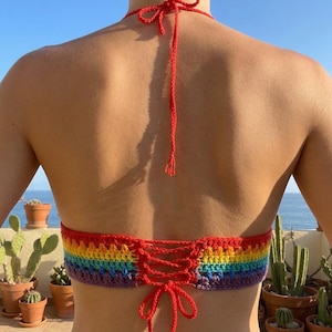 Back view of a handmade pride rainbow crochet bikini top featuring a lace-up corset-style tie closure and thin red halter straps against a sunny outdoor background.