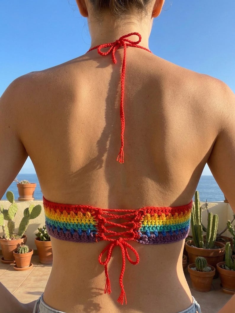 Back view of a handmade pride rainbow crochet bikini top featuring a lace-up corset-style tie closure and thin red halter straps against a sunny outdoor background.