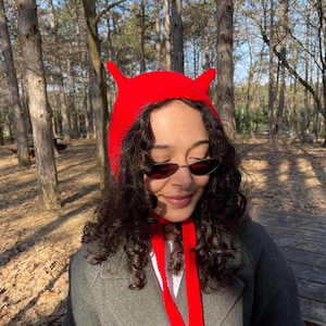 May include: A bright red knit hat with two pointed horns. The hat has a matching red tie. The person is wearing a gray coat and sunglasses. The background is a forest with tall trees.