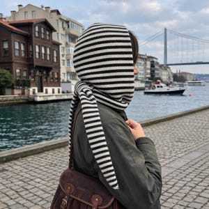 May include: A knitted hood featuring a horizontal black and white striped pattern. The hood is designed to cover the head and neck, with two long, pointed ends that hang down from the chin, allowing for a tie closure under the neck area.