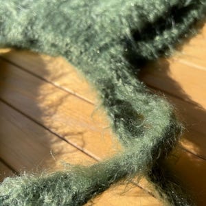 A soft, green fuzzy knitted bonnet lying on a light-colored wooden table. The bonnet features long matching ties and a fluffy, mohair-style texture. Bright, natural sunlight streams across the scene, highlighting the delicate fibers of the knitwear.