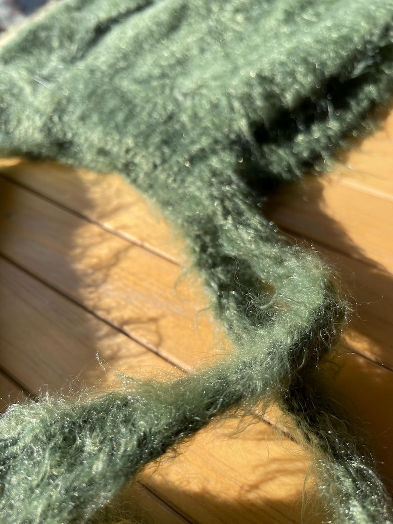 A soft, green fuzzy knitted bonnet lying on a light-colored wooden table. The bonnet features long matching ties and a fluffy, mohair-style texture. Bright, natural sunlight streams across the scene, highlighting the delicate fibers of the knitwear.