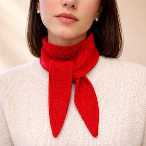 Op de afbeelding: Een felrode gebreide sjaal, om de nek geknoopt. De sjaal is gemaakt van een textuurrijk materiaal en heeft lange, puntige uiteinden. De persoon draagt een crèmekleurige trui. De sjaal voegt een vleugje kleur toe aan de outfit.