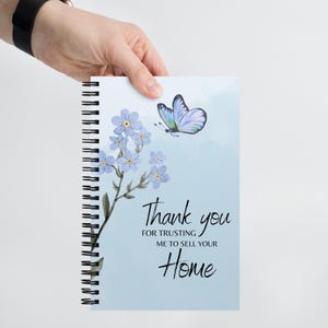 May include: A blue spiral notebook with a watercolor design of forget-me-not flowers and a butterfly. The notebook has the text "Thank you for trusting me to sell your Home".