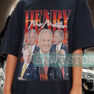 May include: Black t-shirt featuring a graphic of a man's face and the text "HENRY McMaster" in red and gold. The shirt is worn by a person. The background has a mountain and space theme.