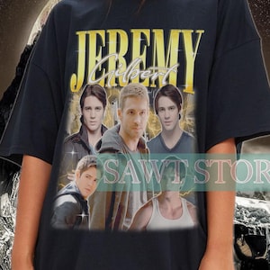 May include: Black t-shirt with the name "Jeremy Gilbert" in gold lettering and a photo of four people. The shirt is worn by a person. The background has a space-themed design.