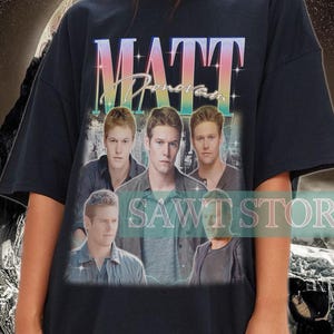 May include: Black t-shirt with a group of men and the text "MATT" in a colourful, gradient font. The shirt has a vintage, slightly faded look. The image is set against a backdrop of a mountain range and a swirling, abstract design.