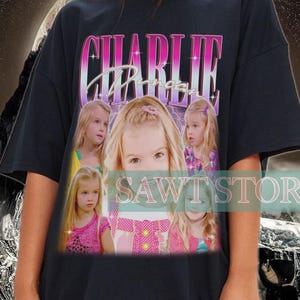 Puede incluir: Camiseta negra de gran tamaño con el nombre "CHARLIE" en texto rosa y blanco. La camiseta presenta un collage de imágenes de una niña. El fondo tiene un diseño retro con un tema de montaña y espacio.