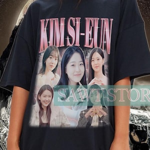 May include: Black t-shirt featuring the name "KIM SI-EUN" in pink lettering above a collage of images. The shirt is worn by a person in front of a surreal background with a mountain and swirling patterns.