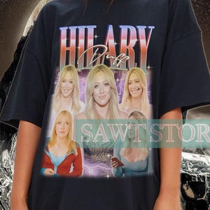 Op de afbeelding: Zwart T-shirt met een vintage afbeelding van Hilary Duff en de tekst "HILARY Duff". Het ontwerp bevat meerdere afbeeldingen van Hilary Duff. Het shirt heeft een ronde hals en korte mouwen. De tekst "SAWI STORE" is ook zichtbaar.