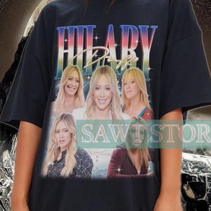 Könnte beinhalten: Schwarzes Oversize-T-Shirt mit einem Grafikdruck von Hilary Duff und dem Text "Hilary Duff" in einem Regenbogen-Verlauf. Die Grafik enthält auch eine schneebedeckte Berglandschaft im Hintergrund.