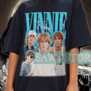 May include: Black t-shirt featuring the name "VINNIE Hacker" in teal block letters above a photo collage of young men. The shirt is worn by a person. The background has a black and teal wavy design.