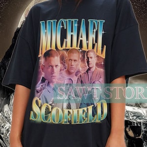 May include: Black t-shirt featuring the name "MICHAEL SCOFIELD" in a retro font, with a faded image of three men. The shirt has a vintage aesthetic, with a background of a mountain range and a starry sky.
