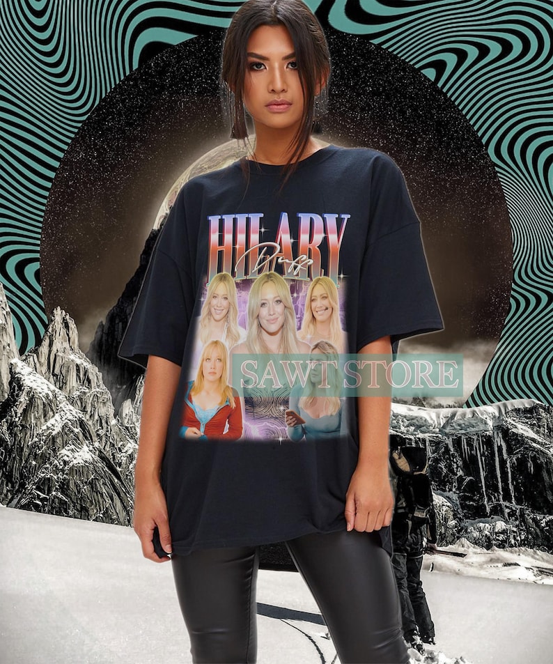 May include: Black t-shirt with a vintage-style graphic of Hilary Duff and the text "HILARY Duff". The design includes multiple images of Hilary Duff. The shirt has a crew neck and short sleeves. The text "SAWI STORE" is also visible.