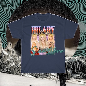 May include: Navy blue t-shirt with a retro design featuring the name "HILARY DUFF". The shirt includes multiple images of Hilary Duff and the text "Scents Store".