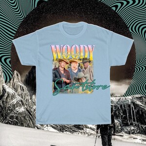 May include: Light blue t-shirt with a vintage-style graphic of four men wearing cowboy hats and the text "Woody's Sanstore".