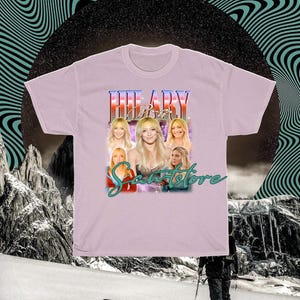 May include: A lilac t-shirt with a graphic of a woman, the name "HILARY" above her, and the text "Sweetstore" below. The graphic includes multiple images of the woman. The background has a black circle and a teal wavy design.