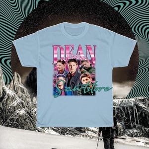 May include: Light blue t-shirt with a graphic print of Dean Winchester from the TV show Supernatural. The graphic features Dean and other characters from the show, with the text "Dean Winchester" and "Samstore" in a retro font.