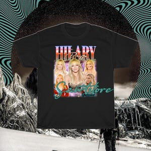 May include: Black t-shirt with a retro design. The design features the text "HILARY Duff" in a stylized font, along with multiple images of a blonde woman. The word "Scantstore" is written below in a script font.