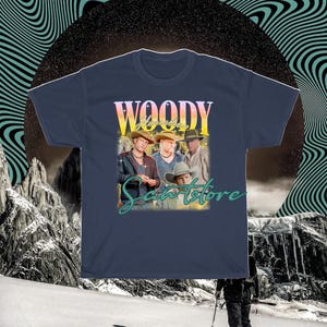 May include: A navy blue t-shirt with a vintage-style graphic of a band called "Woody & The Saints" with the text "Woody & The Saints" in a yellow and orange gradient. The band members are wearing cowboy hats and are standing in front of a sunset background.