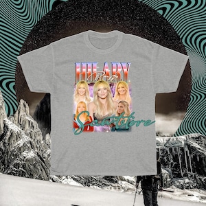 May include: A grey t-shirt with a retro design featuring the name "HILARY" in a colourful, stylised font. The shirt includes multiple images of a woman and the text "Scents Store" in a script font.