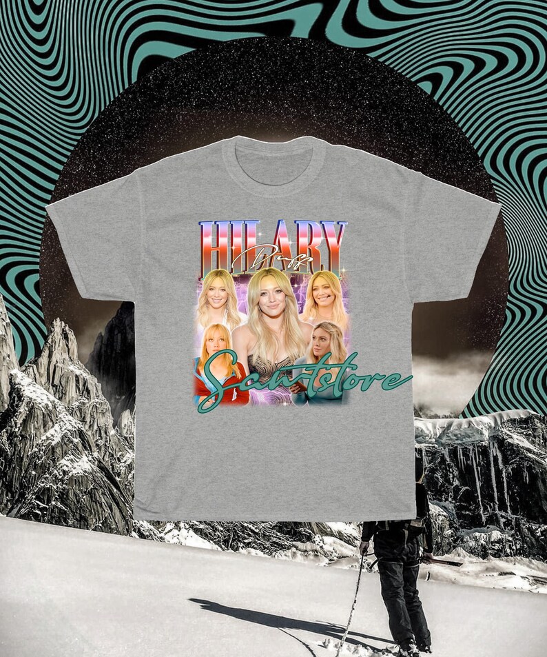 May include: A grey t-shirt with a retro design featuring the name "HILARY" in a colourful, stylised font. The shirt includes multiple images of a woman and the text "Scents Store" in a script font.