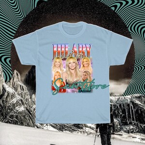 May include: Pale blue t-shirt with a graphic of a woman's image and the name "HILARY" in a retro font. The shirt also features the text "Scents Store" in a cursive font. The background has a retro design.